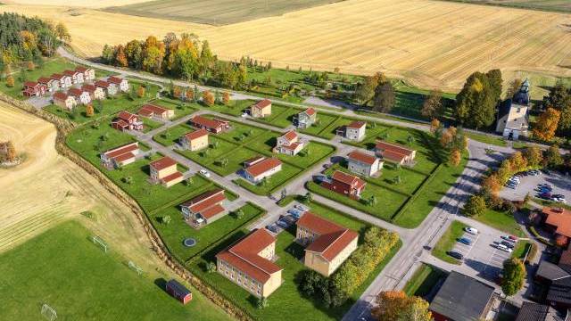 Flygfoto över ett litet bostadsområde med hus med röda tak, omgivet av fält och vägar på landsbygden.