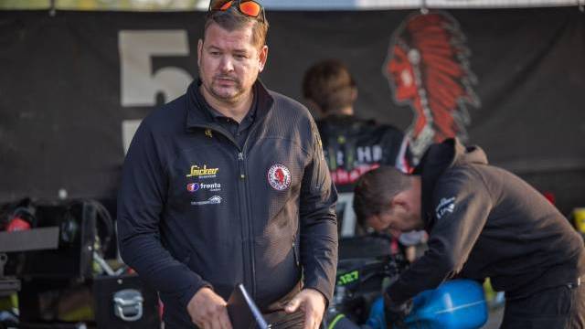 En man i svart jacka håller en anteckningsbok i ett garage, medan en annan person arbetar med utrustning i bakgrunden.