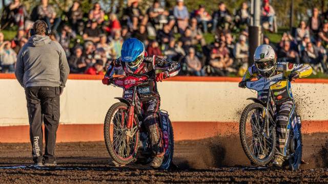 Två motocrossförare accelererar på en grusbana medan en person står i närheten; åskådare tittar på från läktaren i bakgrunden.