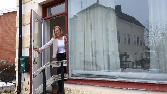 En kvinna står i en öppen dörröppning på en liten veranda utanför en byggnad med stora fönster, håller dörren öppen och tittar framåt.