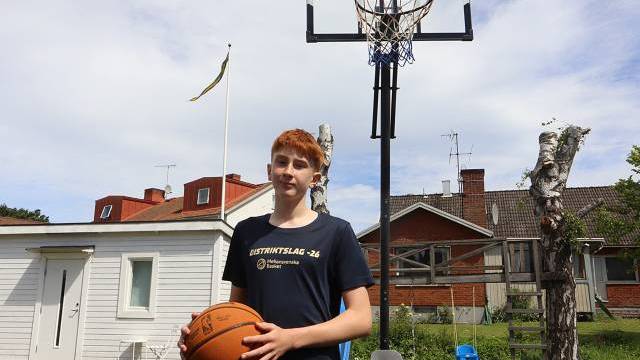 En tonårspojke som håller en basketboll står framför en utomhusbasketkorg i en trädgård, med ett vitt skjul och hus av rött tegel i bakgrunden.