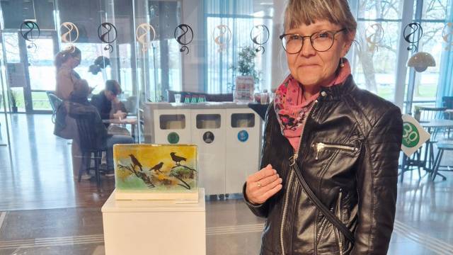 En kvinna i svart skinnjacka står bredvid en liten fågelmålning på en piedestal i en modern inomhusmiljö.