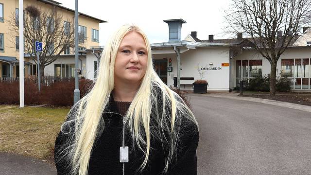 En kvinna med långt blont hår och svart jacka står utomhus framför en byggnad märkt 