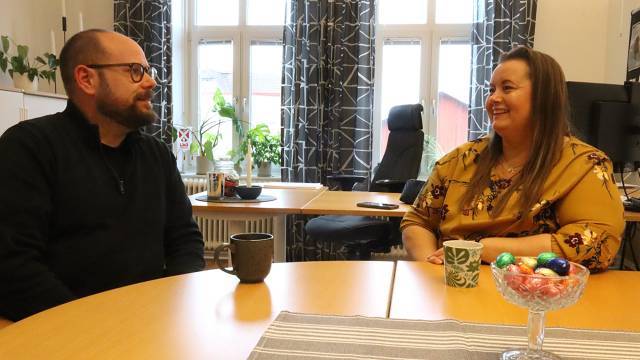 Två personer sitter vid ett runt bord på ett kontor och ler och pratar, med muggar och en skål med godis på bordet.