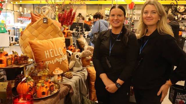 Två kvinnor står bredvid en utställning med höstinspirerade dekorationer, bland annat pumpor, ljus och en kudde med texten ”Hello Pumpkin”, inne i en butik.
