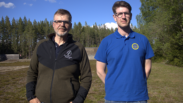 Två män står utomhus på en gräsmatta med träd och en blå himmel i bakgrunden; den ene bär en mörk jacka, den andre en blå polotröja med en cirkelformad logotyp.
