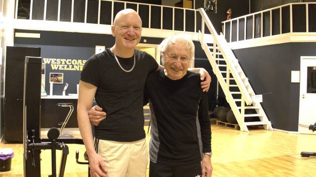 Två äldre män i träningskläder står sida vid sida och ler och poserar i ett gym med trägolv och träningsutrustning.