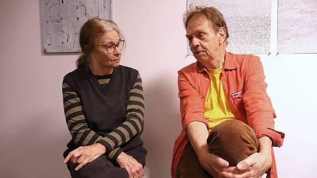 Två äldre vuxna sitter inomhus, vända mot varandra och samtalar. Kvinnan bär glasögon, randig skjorta och mörk väst, mannen har en orange skjorta över en gul T-shirt. På väggen hänger en konst.