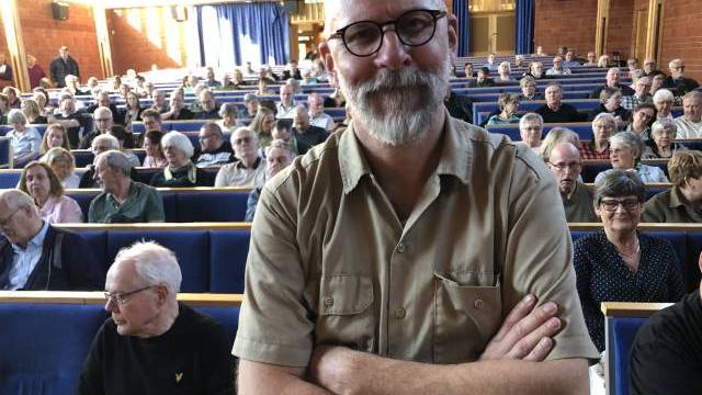 En man med glasögon och skägg står med armarna i kors framför en publik som sitter i ett auditorium av trä.