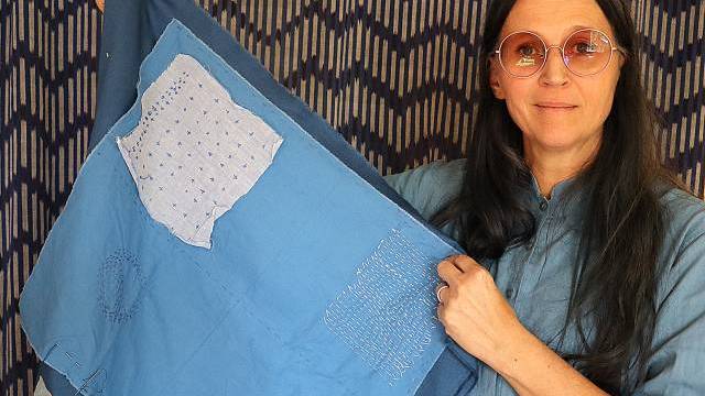 En person med runda glasögon och blå skjorta håller upp en blå textil med synliga lappar och sömmar mot en bakgrund av mönstrat tyg.