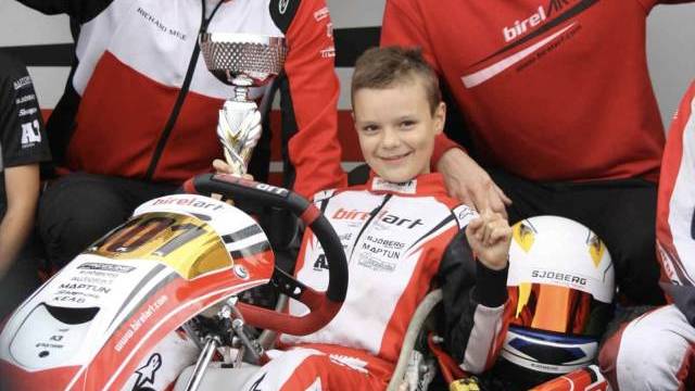 En ung gokartförare sitter i en racingkart och håller i en pokal, flankerad av två leende vuxna i racingteamdräkt; en annan pokal hålls i bakgrunden.