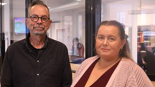 Två vuxna står inomhus i en kontorsmiljö och tittar in i kameran. Mannen bär glasögon och en mörk skjorta; kvinnan bär en rosa kofta och halsband.
