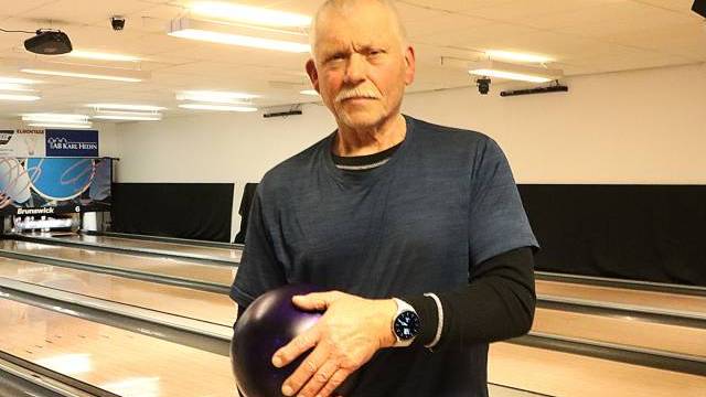 En äldre man står i en bowlinghall och håller i ett lila bowlingklot, klädd i en blå t-shirt, svarta byxor och en klocka. Bowlingbanor och käglor syns i bakgrunden.