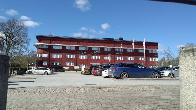 En röd trevåningsbyggnad med flera vita flaggor framför, en parkeringsplats full av bilar och en klarblå himmel i bakgrunden.