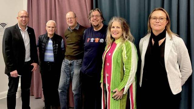 Sex vuxna står sida vid sida framför mörka och ljusa gardiner och poserar för ett gruppfoto. Fyra män står till vänster och två kvinnor till höger.