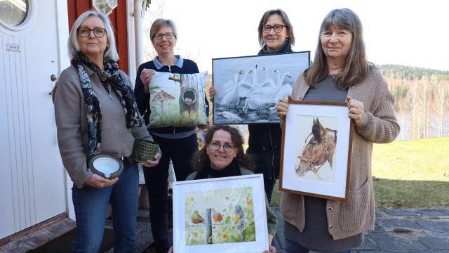 Fem kvinnor står utomhus vid ett hus, var och en med ett konstverk i handen, bland annat målningar av fåglar, en häst och en naturscen. I bakgrunden syns träd och ett vattendrag.