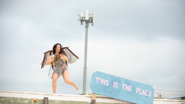En utklädd person står barfota på ett busstak nära en blå skylt med texten 