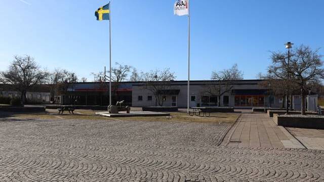 Ett asfalterat torg med två flaggstänger, en med en svensk flagga och en med en vit flagga, framför en låg byggnad under en klarblå himmel.