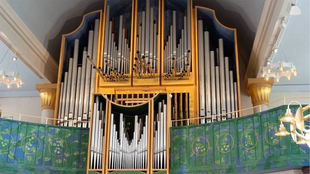 Stor piporgel med metallpipor och en dekorativ balkong i grönt och guld i en väl upplyst kyrkorum.