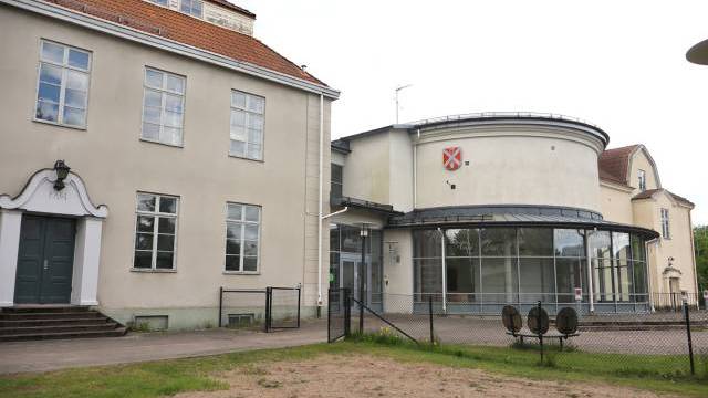 En beige tvåvåningsbyggnad med stora fönster och en modern glasutbyggnad, omgiven av ett stängsel av trådnät och en grusplan i förgrunden.