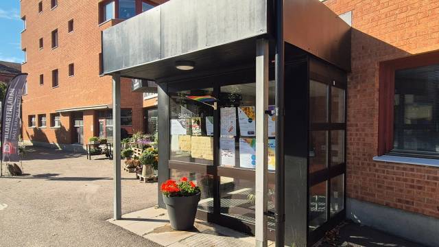 En ingång med glasparti och ett tak av metall, en anslagstavla täckt av affischer och en svart blomkruka med röda och gula blommor utanför en tegelbyggnad en solig dag.