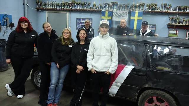 En grupp människor står bredvid en svart och rosa racerbil i ett garage, med pokaler på hyllor och svenska flaggor på väggen bakom dem.