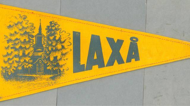 Gul vimpel med en illustration av en kyrka och träd till vänster och ordet ”LAXÅ” i fetstil till höger.