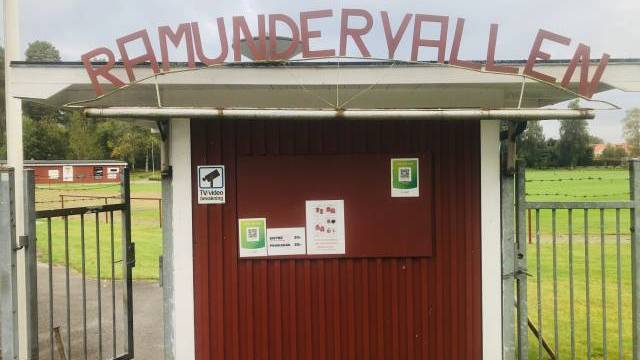 Entrégrind vid Ramundervallens idrottsplats med ett rött skjul, QR-kodskyltar och meddelande om videoövervakning. Gräsplan och träd syns i bakgrunden.
