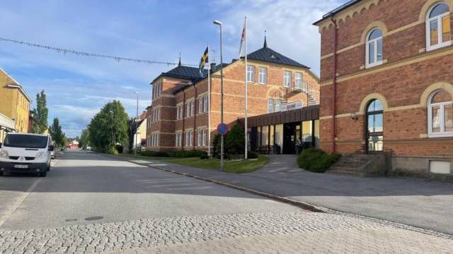 En byggnad i rött tegel med välvda fönster, omgiven av träd och flaggor. En skåpbil står parkerad på gatan och himlen är delvis molnig.