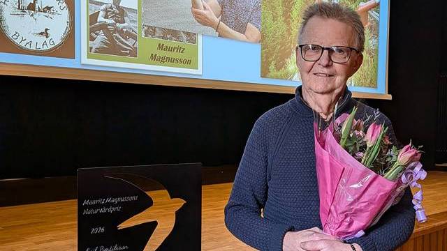 En äldre person som håller i en bukett rosa blommor står vid en prisplakett, med en presentationsbild med foton och namn i bakgrunden.