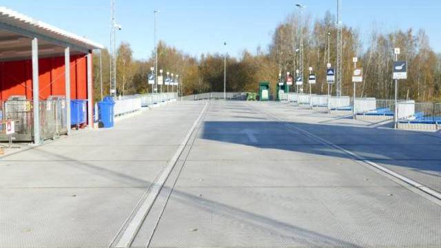 En ren, tom återvinningscentral utomhus med märkta soptunnor och skyltar, omgiven av träd och metallstaket under en klar himmel.