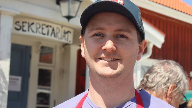 En man med svart keps och rött förkläde står ute i solskenet och ler mot kameran. I bakgrunden syns en byggnad med en skylt där det står ”SEKRETARIAT”.