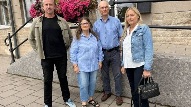 Fyra vuxna står utanför en byggnad med en blomlåda, klädda i vardagskläder, och tittar in i kameran.