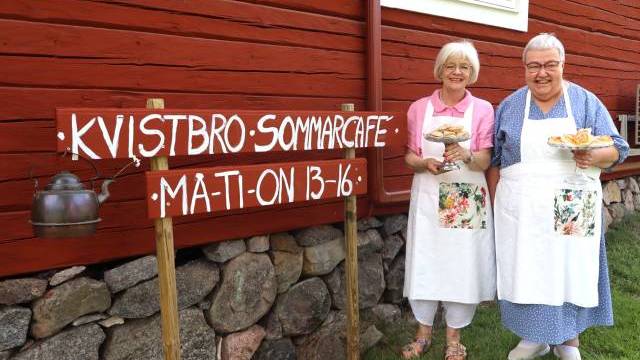 Två kvinnor i förkläden står vid en röd träbyggnad och håller i tallrikar med bakverk bredvid en skylt där det står ”KVISTBRO SOMMARCAFE MAT-ON 13–16”, med en vattenkokare hängande på skylten.