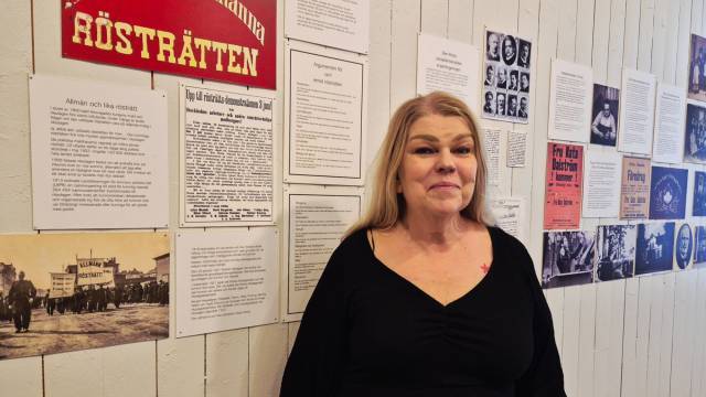En kvinna i svart topp står framför en vägg med historiska dokument, fotografier och affischer, bland annat en med svensk text om rösträtt.