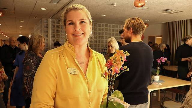 En kvinna i gul blus med en orkidé i handen står leende i ett rum med flera personer samlade i bakgrunden.