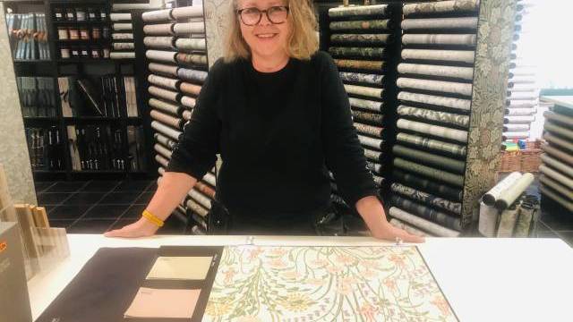 En person står bakom en disk och visar tapetprover och färgprover i en butik med hyllor med tapetrullar i bakgrunden.