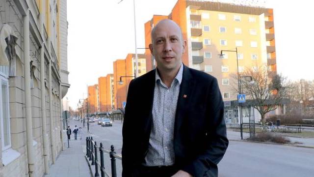 En man i kostym står på en trottoar i staden med flerbostadshus och några fotgängare i bakgrunden i dagsljus.