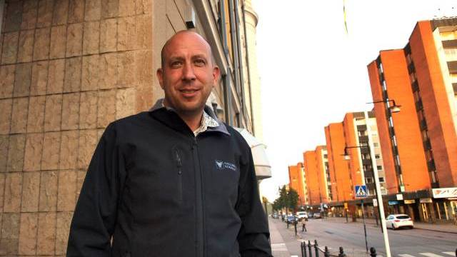 En man i mörk jacka står på en trottoar i en stad bredvid en byggnad, med orangefärgade flerfamiljshus och en gata i bakgrunden.