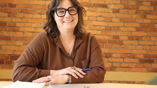 En person med glasögon sitter vid ett bord med en bärbar dator, anteckningsbok och penna och ler mot en bakgrund av en tegelvägg.