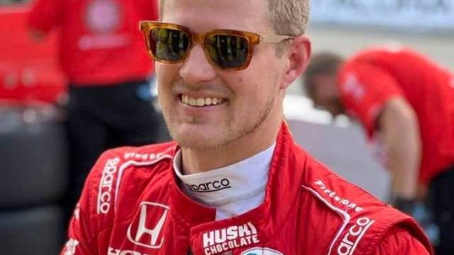 En racerbilsförare i röd racingdräkt och solglasögon ler när han står utomhus tillsammans med andra teammedlemmar i matchande röda kläder.