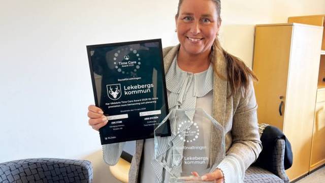 En kvinna står inomhus och ler, med ett inramat certifikat i ena handen och ett pris i klarglas i den andra, båda med texten 