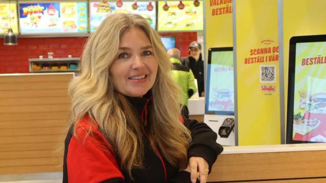 En kvinna med långt blont hår och en svart-röd jacka står inne på en snabbmatsrestaurang nära självbeställningsskärmar och menytavlor.