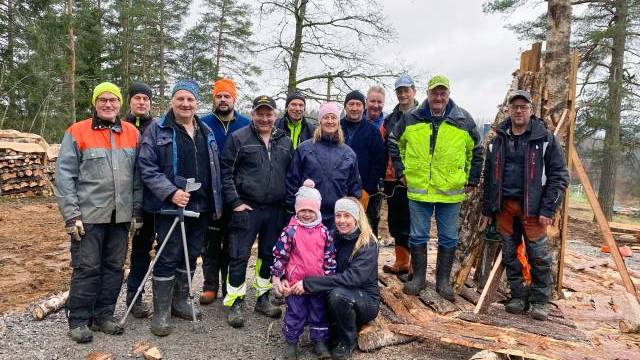 En grupp människor, däribland två barn, står utomhus i arbetskläder och skyddsutrustning vid en avverkningsplats som omges av avverkat virke och träd.