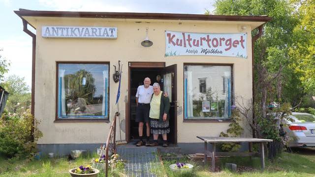 Två personer står i dörröppningen till en liten byggnad med skyltar med texten 