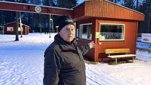 En äldre man i svart jacka och mössa står i snön och pekar mot en röd träbyggnad med en bänk, som ligger i ett skogsområde med andra byggnader i närheten.