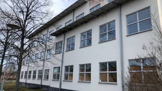 En vit byggnad i tre våningar med flera fönster, ett platt tak och synlig väderbiten skyltning på den övre fasaden; lövlösa träd och gräs i förgrunden.