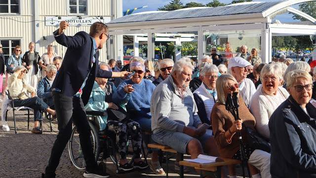 En man står upp och gestikulerar livligt mot en sittande publik bestående främst av äldre vuxna vid ett utomhusevenemang en solig dag.
