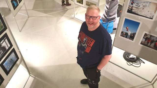 En man med glasögon och skägg står och ler i ett konstgalleri, omgiven av fotoutställningar på vita paneler.