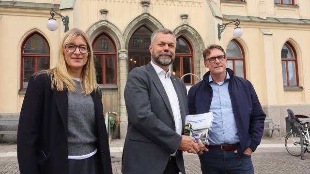 Tre vuxna står utanför en historisk byggnad; en man i kostym håller i några dokument, med en kvinna och en annan man på vardera sidan om sig.